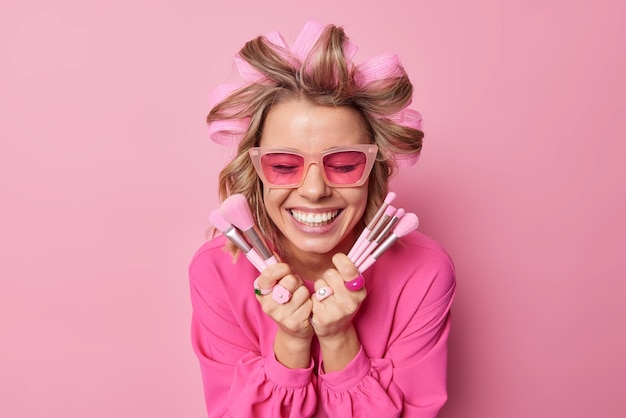 Gelukkige dame past haarrollers toe voor het maken van kapsel draagt trendy zonnebril zand blouse houdt cosmetische borstels die make-up gaan aanbrengen glimlacht breed geïsoleerd over roze muur Vrouwen schoonheidsbehandelingen