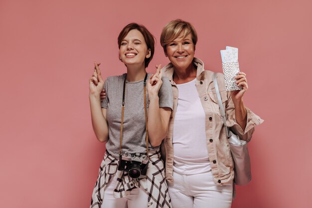 Gelukkige dames met kort kapsel en charmante glimlach in trendy outfit knuffelen, kruising vinger en kaartjes op roze achtergrond te houden.