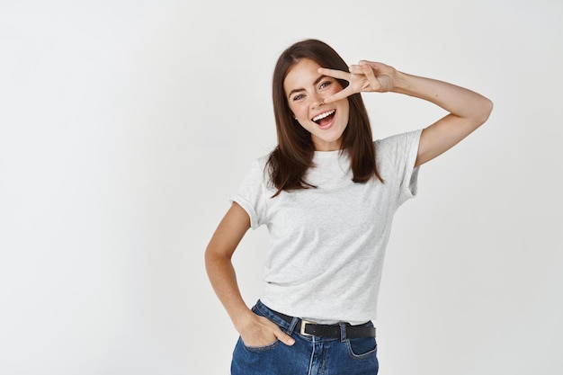 Gelukkige donkerbruine vrouw die vrede of overwinningsteken op oog toont en glimlacht, vreugde en opwinding uitdrukt, die zich in t-shirt tegen witte muur bevindt