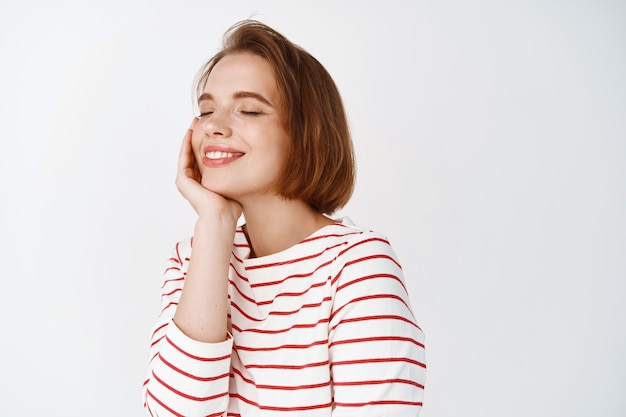 Gelukkige emoties. Mooie vrouw met kort haar en natuurlijk fris gezicht, ogen dicht en glimlachen, dromend over iets moois, staande tegen een witte muur