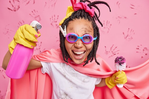 Gratis foto gelukkige emotionele vrouw die superheld schoonmaakt, houdt chemisch spuitmiddel vast en vuile borstel helpt je met huishoudelijk werk draagt een veiligheidsbril die over roze muur wordt geïsoleerd
