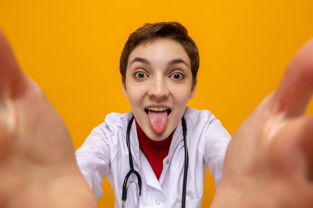 Gelukkige en grappige jonge vrouwelijke arts in witte jas met stethoscoop om nek die selfie neemt die tong uitsteekt op oranje