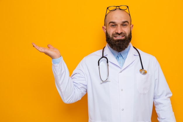 Gelukkige en positieve bebaarde man arts in witte jas met stethoscoop om nek met bril op zijn hoofd op zoek naar presentatie van kopieerruimte met arm van zijn hand