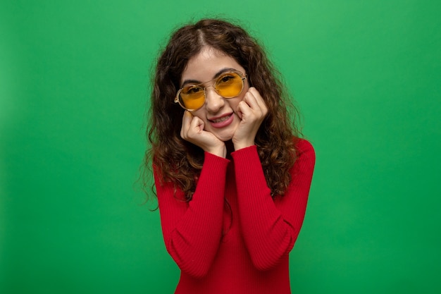 Gelukkige en positieve jonge mooie vrouw in rode coltrui met een gele bril die vrolijk glimlacht met handen op haar wang die over groene muur staat