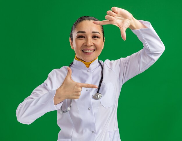 Gelukkige en positieve jonge vrouwelijke arts in een witte medicijnjas met een stethoscoop om de nek die een framegebaar maakt met vingers die over de groene muur staan