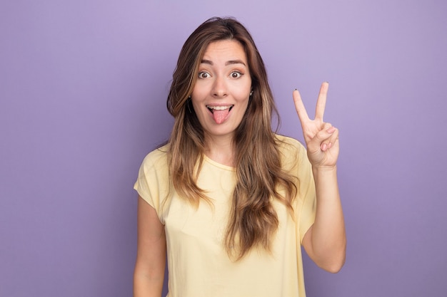 Gelukkige en vrolijke jonge mooie vrouw in beige t-shirt kijkend naar camera die tong uitsteekt met v-teken dat over paarse achtergrond staat