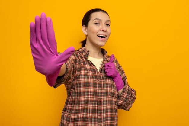 Gelukkige en vrolijke jonge schoonmaakster in geruit hemd in rubberen handschoenen die vijfde toont met palm, high five die op oranje staat