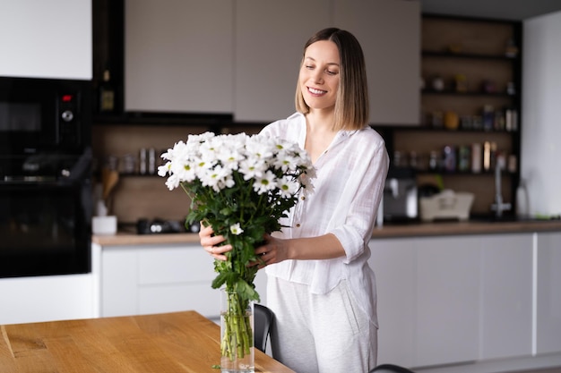 Gelukkige en vrolijke jonge vrouw in het wit die witte bloemen schikt thuis in de keuken