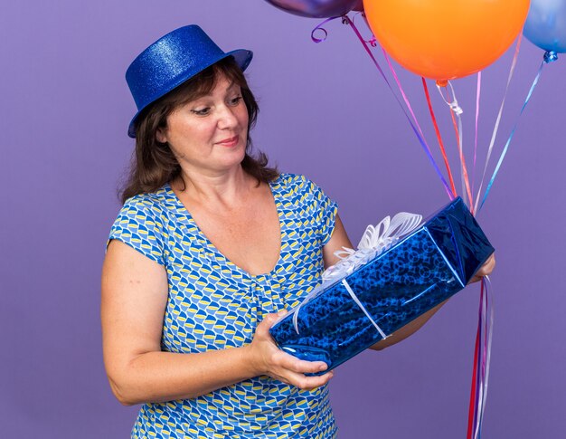 Gelukkige en vrolijke vrouw van middelbare leeftijd in feestmuts die een stel kleurrijke ballonnen vasthoudt en ernaar kijkt met een glimlach die een verjaardagsfeestje viert dat over de paarse muur staat