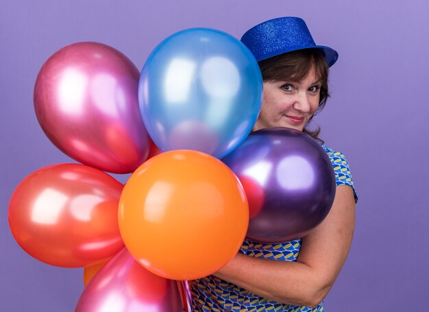 Gelukkige en vrolijke vrouw van middelbare leeftijd in feestmuts met een bos kleurrijke ballonnen die lacht om een verjaardagsfeestje te vieren dat over een paarse muur staat