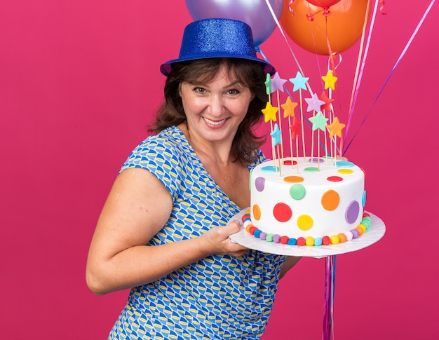 Gelukkige en vrolijke vrouw van middelbare leeftijd in feestmuts met kleurrijke ballonnen met verjaardagstaart die breed glimlacht
