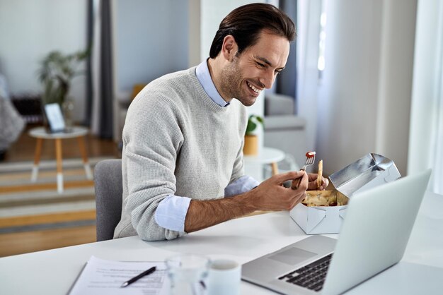 Gelukkige freelance werknemer aan het eten terwijl hij thuis op een computer op het internet surft