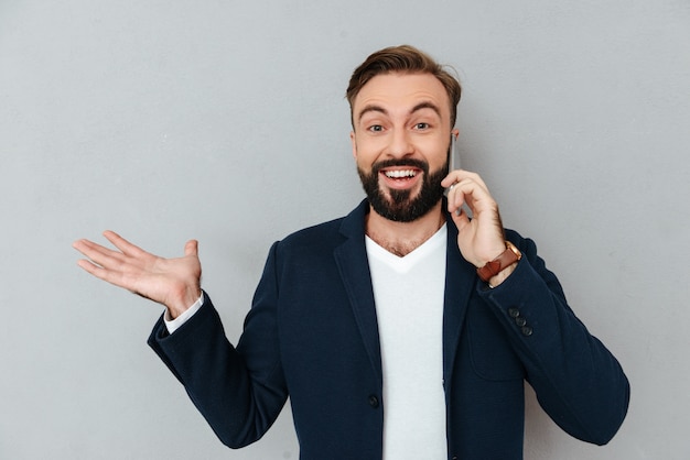 Gelukkige gebaarde mens in bedrijfs door smartphone spreken en kleren die copyspace op het pond een gat maken in terwijl het bekijken de camera over grijs