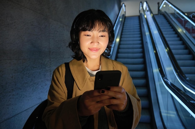 Gelukkige glimlachende jonge vrouw die zich op roltrap bevindt die smartphone in beide handen houdt die babbelt