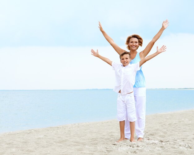 Gelukkige glimlachende moeder en zoon die zich op het strand met opgeheven handen bevinden.