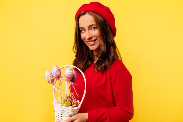 Gelukkige glimlachende mooie vrouw met krullend haar die rode trui en rode baret dragen die met Pasen-mand stellen over geïsoleerde gele muur