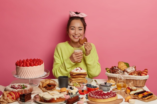 Gelukkige huisvrouw bakte veel smakelijke desserts, wacht op echtgenoot, heeft heerlijk ontbijt, eet haverkoekjes met melk, poseert binnen.