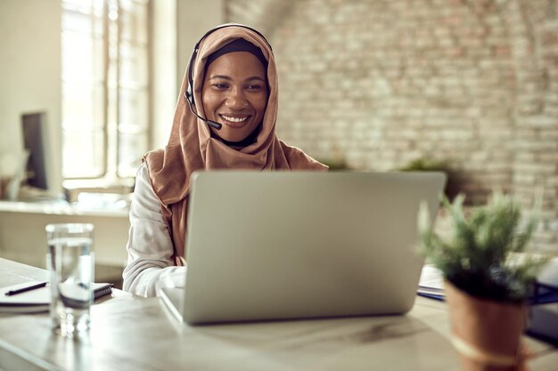 Gelukkige islamitische zakenvrouw die op een computer werkt en communiceert via een headset op kantoor