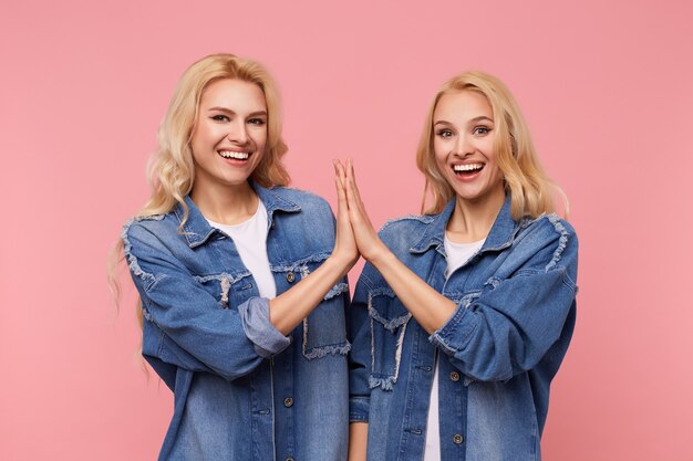Gratis foto gelukkige jonge aantrekkelijke langharige blonde zusters klappen opgeheven handpalmen van elkaar en kijken met een brede glimlach naar de camera, staande over roze achtergrond