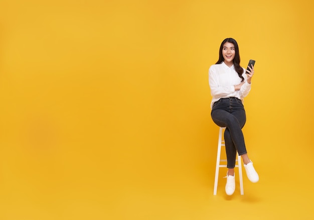 Gelukkige jonge Aziatische vrouw die mobiele telefoon toont terwijl ze op een witte stoel zit