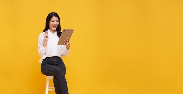 Gelukkige jonge Aziatische vrouw met een glimlach die een tablet gebruikt terwijl ze op een witte stoel zit en kijkt