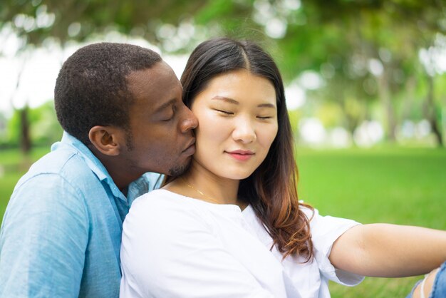 Gelukkige jonge Aziatische vrouw met gesloten ogen die kus van Afrikaanse vriend in openlucht voelen