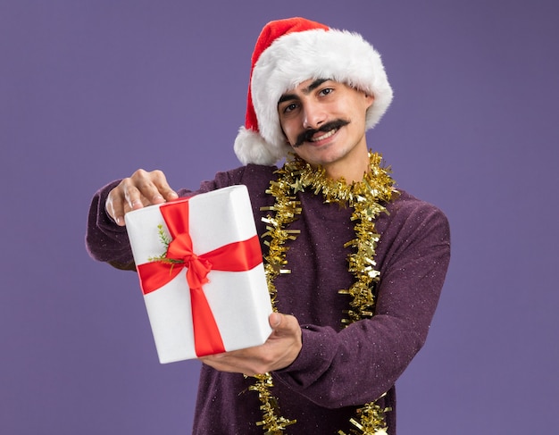 Gelukkige jonge besnorde man met een kerstmuts met klatergoud om zijn nek en een kerstcadeau met een glimlach op het gezicht die over een paarse muur staat