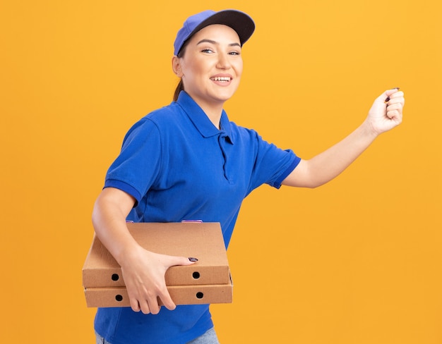 Gelukkige jonge bezorger in blauw uniform en pet haast zich rennen voor het bezorgen van pizzadozen voor klant over oranje muur