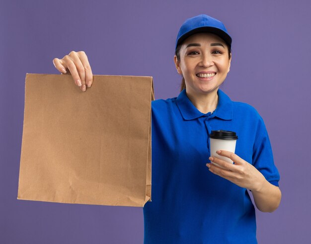 Gelukkige jonge bezorger in blauw uniform en pet met papieren pakket en papieren beker met een glimlach op het gezicht over een paarse muur