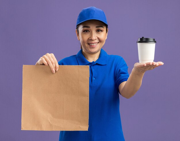 Gelukkige jonge bezorger in blauw uniform en pet met papieren pakket met papieren beker met glimlach op gezicht over paarse muur