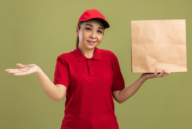 Gelukkige jonge bezorger in rood uniform en pet met papieren pakket met glimlach op gezicht presenterend met arm over groene muur