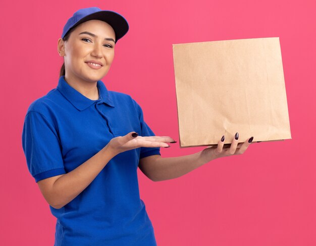 Gelukkige jonge bezorgvrouw in blauw uniform en pet met papieren pakket presenteren met armen kijken naar voorkant glimlachend vrolijk staande over roze muur