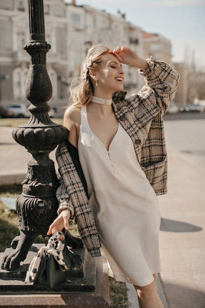 Gratis foto gelukkige jonge blonde vrouw in trendy witte jurk en tweed geruite jas glimlacht oprecht en poseert op het stadsplein