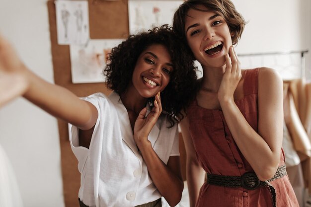 Gelukkige jonge dames in stijlvolle outfits poseren in kantoor van modeontwerper Charmante vrouw in linnen jurk en donkerhuidige dame in witte blouse nemen selfie