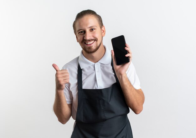 Gelukkige jonge knappe kapper die eenvormig draagt die mobiele telefoon en duim toont die omhoog op wit met exemplaarruimte wordt geïsoleerd