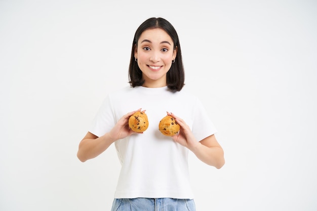 Gelukkige jonge koreaanse vrouw die twee cupcakes over de borst toont die glimlacht en kijkt naar gelukkig gebak en bakkerij