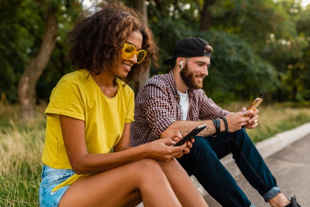 Gelukkige jonge lachende vrienden zitten park met behulp van smartphones, man en vrouw samen plezier