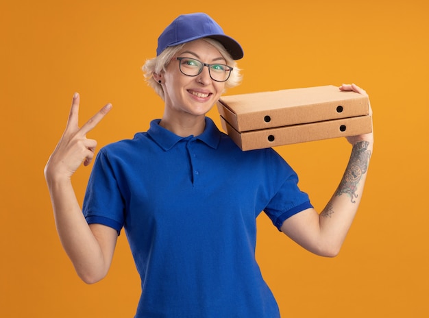 Gelukkige jonge leveringsvrouw in blauw uniform en GLB die glazen dragen die pizzadozen glimlachen die v-teken tonen over oranje muur