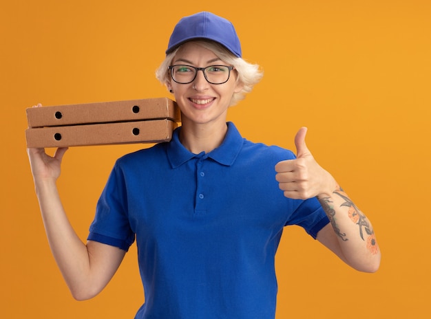 Gelukkige jonge leveringsvrouw in blauw uniform en GLB die glazen dragen die pizzadozen glimlachend tonen duimen over oranje muur houden