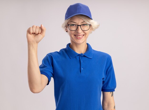 Gelukkige jonge leveringsvrouw in blauw uniform en GLB die zelfverzekerd opheffende vuist glimlachen die zich over witte muur bevindt