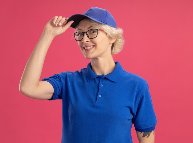 Gelukkige jonge leveringsvrouw in blauw uniform en pet die zelfverzekerd aanraken GLB glimlachend status over roze muur kijken