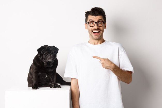 Gelukkige jonge man die zijn schattige hond laat zien, met de vinger naar zwarte mopshond wijst en glimlacht, staande op een witte achtergrond