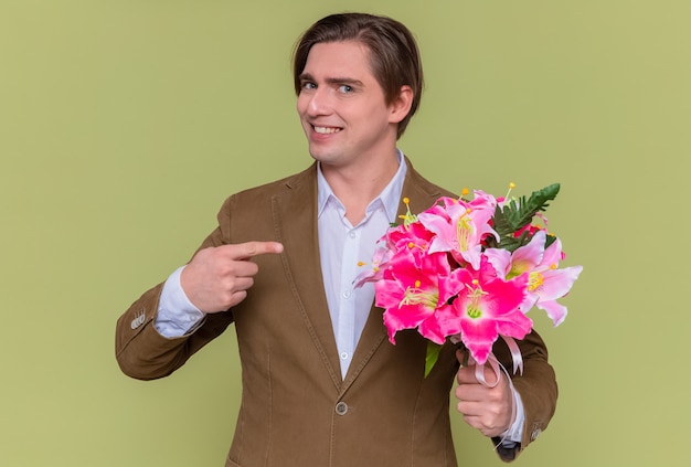 Gelukkige jonge man met boeket bloemen wijzend met wijsvinger naar het glimlachend vrolijk gaat feliciteren met internationale vrouwendag concept staande over groene muur