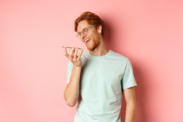 Gelukkige jonge man met rood haar die blij glimlacht terwijl hij een spraakbericht opneemt op een smartphone die praat met...