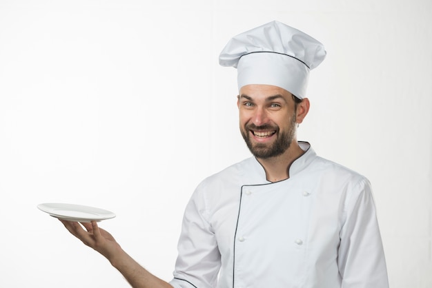 Gelukkige jonge mannelijke chef-kok die een lege witte plaat houdt tegen witte achtergrond