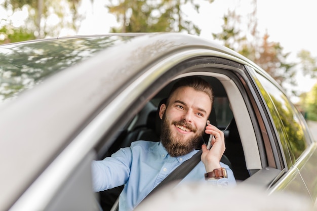 Gelukkige jonge mens die door auto reist die mobiele telefoon met behulp van