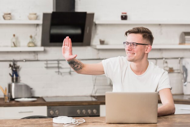 Gelukkige jonge mens die zijn hand met laptop op lijst in de keuken golft