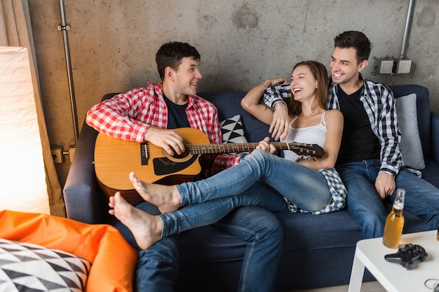 Gelukkige jonge mensen die lol hebben, vrienden feesten thuis, hipster gezelschap samen, twee mannen een vrouw, gitaar spelen, glimlachen, positief, ontspannen, bier drinken, jeans, shirts, casual stijl