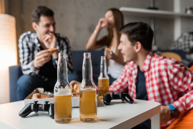 Gelukkige jonge mensen die pizza eten, bier drinken, plezier maken, vriendenfeestje thuis, hipster gezelschap samen, twee mannen één vrouw, glimlachen, positief, ontspannen, rondhangen, lachen,