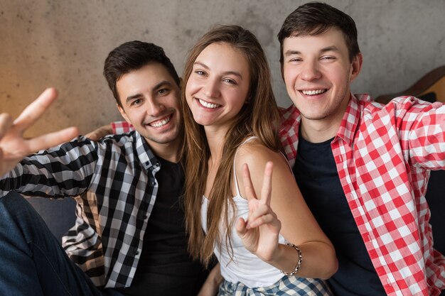 Gelukkige jonge mensen maken grappige selfie foto, zittend op de bank, plezier maken, vrienden thuisfeestje, hipster gezelschap samen, twee mannen een vrouw, jeugdstijl, glimlachen, positieve emotie, in de camera kijken